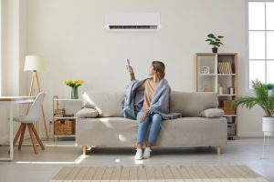 Woman who's sitting on sofa under warm plaid in living room switches off her hvac air conditioner on wall